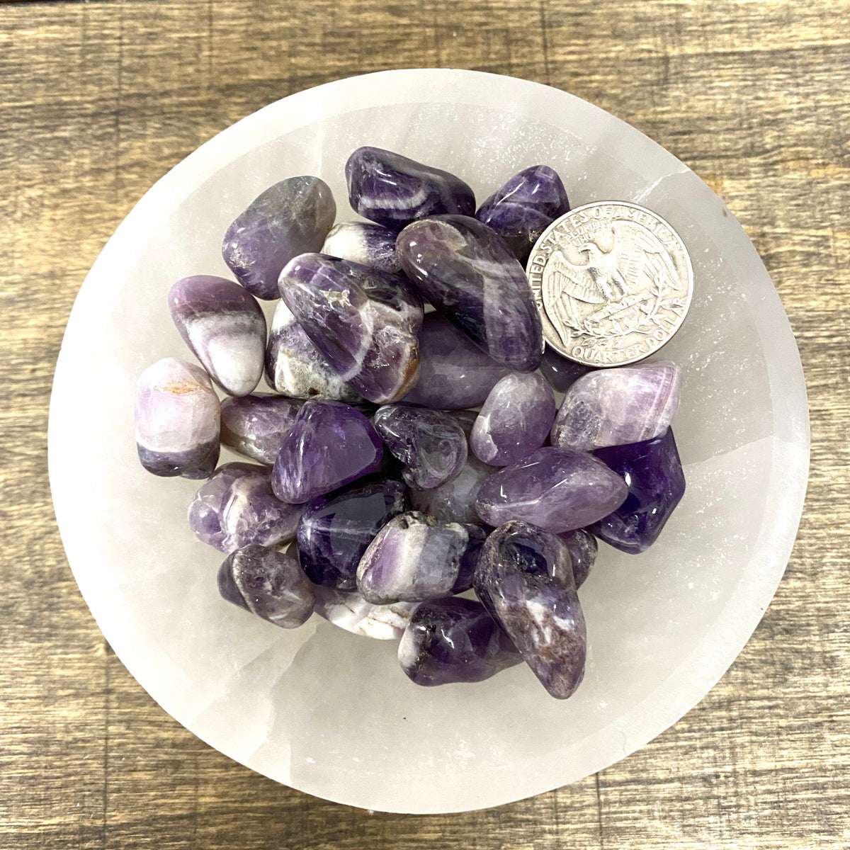 Overhead comparison shot of a US quarter coin and various Chevron Amethyst tumbled stones in a small bowl.