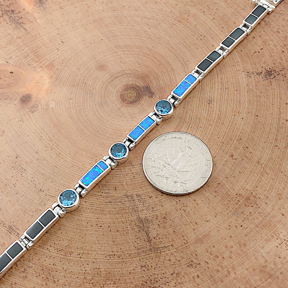 A comparison shot of a US quarter coin and a sterling silver bracelet that is inlaid with Black Jade, Blue Cultured Opal, and is set with three London Blue Topaz stones that is laid flat