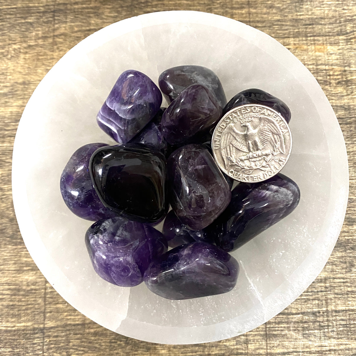 Overhead comparison shot of a US quarter coin and various Amethyst tumbled stones in a small bowl.