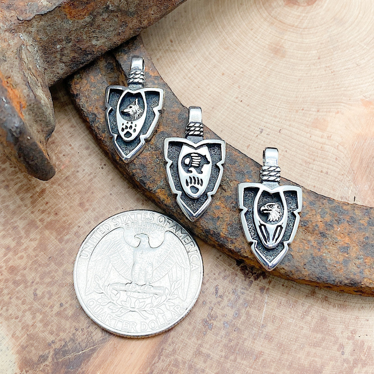 Comparison shot of a US quarter coin various arrowhead pendants laid in a row, each with a different animal and symbol etched within the pendant.